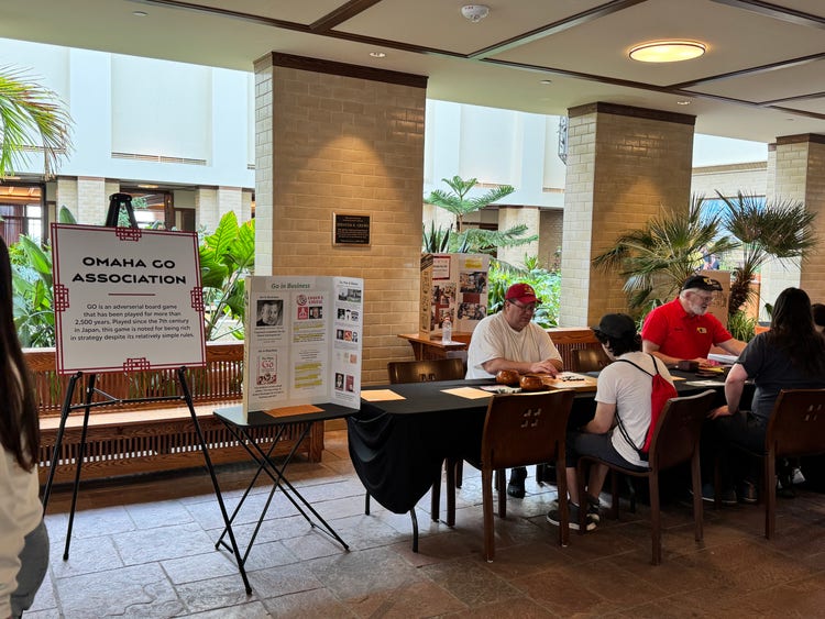 A group of people sits at a table in a spacious indoor area with plants, engaging in conversation. A sign reads