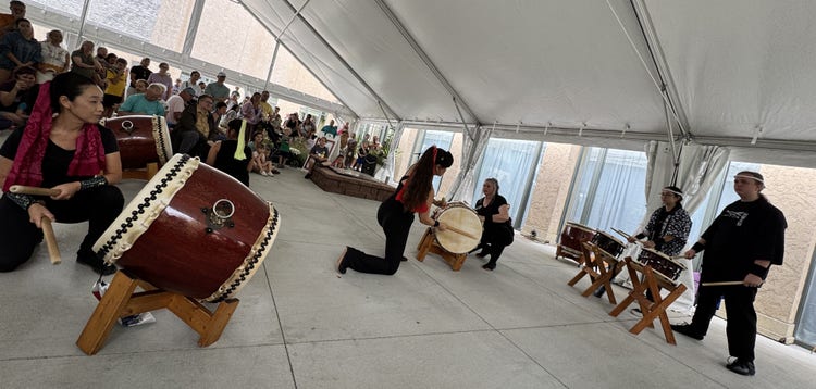 A group of performers is playing traditional drums under a large white tent. The musicians are focused, with one kneeling and playing a large drum, while others stand with smaller drums on stands. An attentive audience watches in the background.