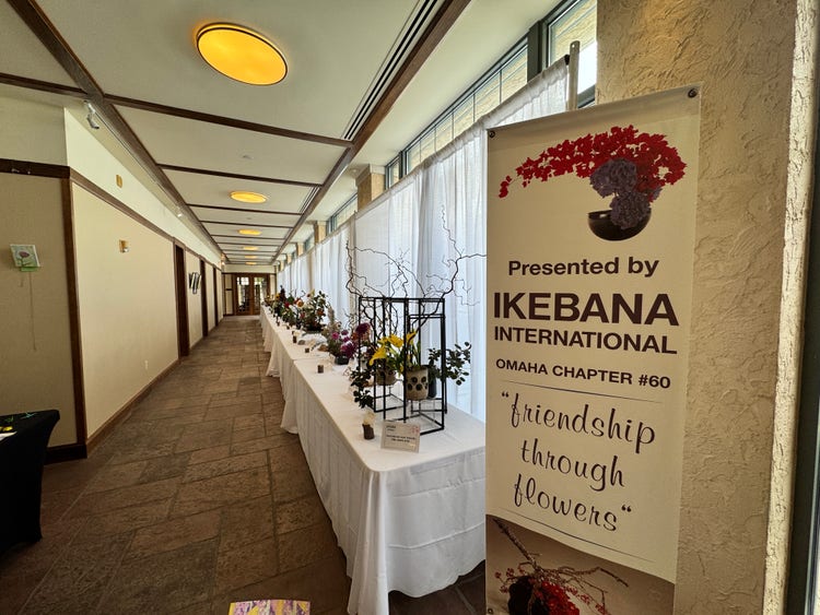 A hallway features a long table adorned with elegant ikebana flower arrangements, each uniquely styled. A banner nearby reads