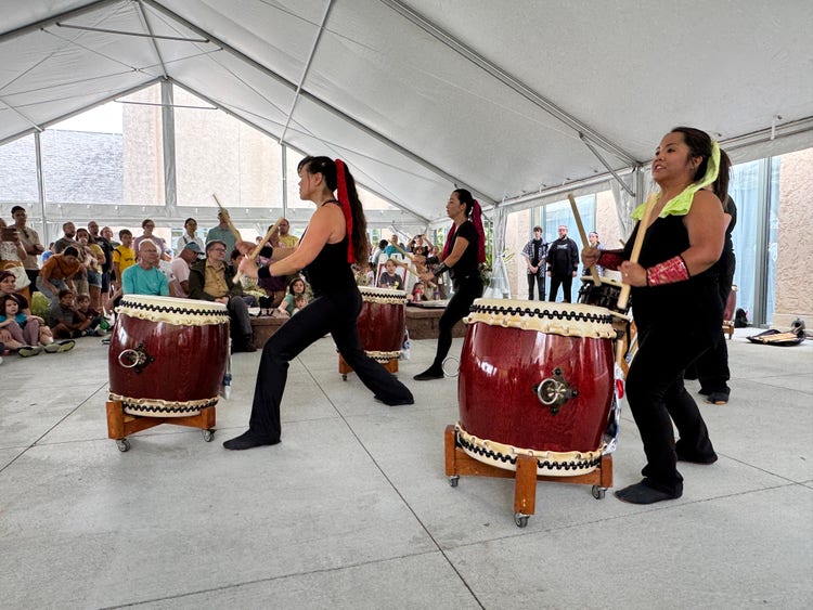 A group of performers energetically play traditional Japanese taiko drums under a large white tent, with three visible drummers in black outfits and colorful accents. An audience sits attentively in the background, watching the lively performance.